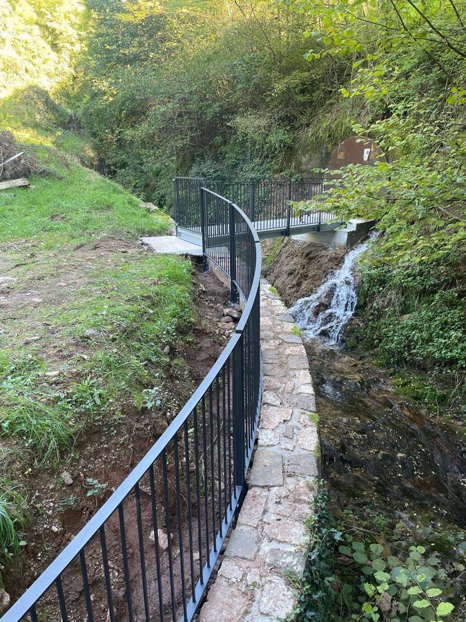 Ringhiera in ferro che segue la curva di un passaggio lungo un ruscello, a Faedo.